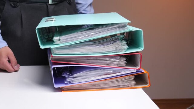Person standing behind a desk, holding a heavy stack of ring binders filled with paperwork, illustrating concepts of office work, administration, and documentation
