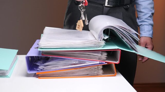 Person standing behind a table full of stacked ring binders and paper documents, holding house keys with a small wooden house charm, representing homeownership bureaucracy and mortgage processes