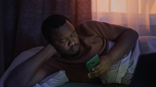 Zoom in shot of African American young man lying in bed at night, scrolling on smartphone with laptop open nearby. Ideal for concepts of digital addiction, insomnia, and sleep hygiene