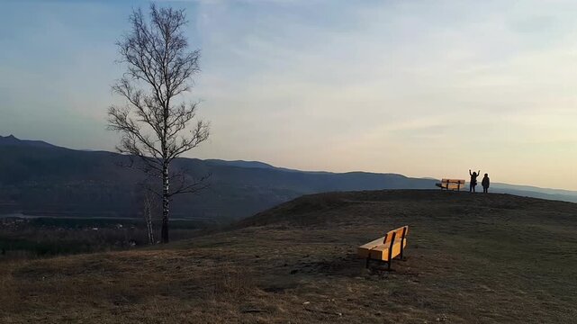 Lone birch tree on a hill with two yellow benches, distant silhouettes of two people raising arms and shouting to the sky, spiritual practice, mountain ridge at cool sunset, deep autumn, dry grass, pa