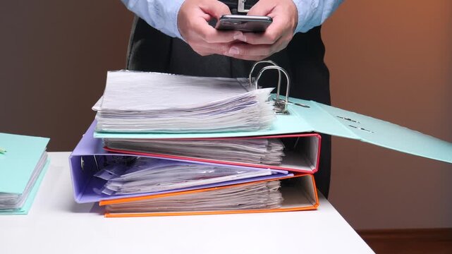 Businessman's hands interacting with a smartphone above a desk stacked with ring binders full of documents. Illustrating the integration of digital communication with traditional record keeping tasks