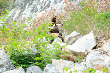 Fototapeta premium Male soldier in camouflage uniform sitting on a rock on the edge high cliff in a mountainous area closely observing the border situation.