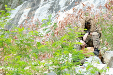 Fototapeta premium Male soldier in camouflage uniform sitting on a rock on the edge high cliff in a mountainous area closely observing the border situation.