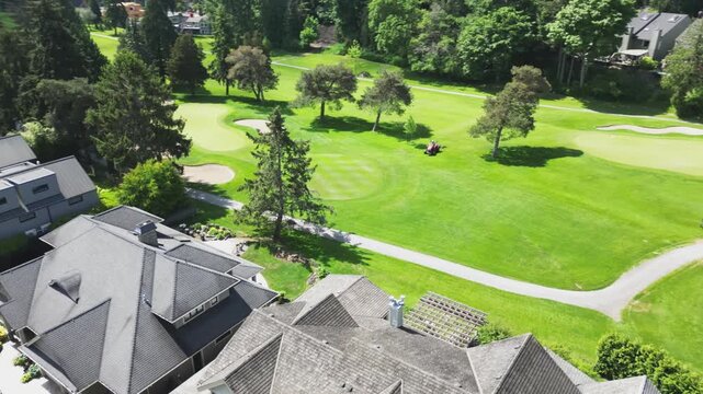 High-angle aerial perspective slowly pans right across the grey shingled rooftops of luxury residential houses to reveal a bright green golf course fairway where a groundskeeper operates a red riding