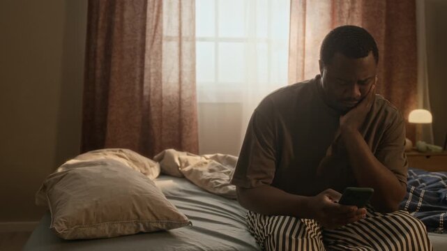 Medium long shot of young African American man sitting on bed, looking at smartphone with sad expression. Perfect for mental health awareness, sleep disorder, or digital fatigue commercial