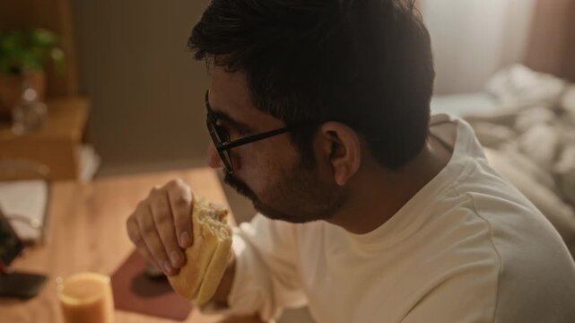 Tilt up shot of Indian young man with glasses eating sandwich while typing on laptop at desk. Ideal for marketing campaigns focusing on remote work, busy lifestyle, multitasking concepts