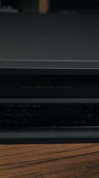 Male hand lifts the plastic flap on a retro black VCR to reveal hidden control buttons. Soft studio lighting highlights the texture of the vintage electronics on a wooden surface.