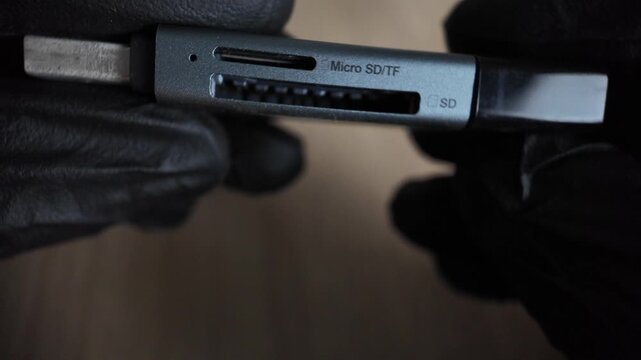 A close up shot of a gray aluminum USB card reader with slots for Micro SD, TF, and SD memory cards held by black gloved hands.
