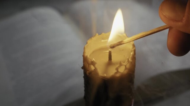 A hand uses a match to light a textured beeswax candle wick with a blurred open book in the background. Concept of historical study, spiritual ritual, magic, and retro education.