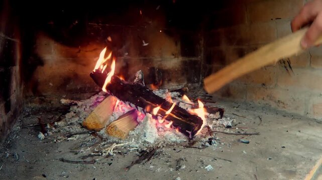 Close-up side view of a brick fireplace interior with flames burning, glowing coals, and a man's hand adding a log to the fire