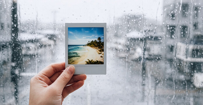 Mano con fotografia istantanea di paesaggio tropicale, sfondo con superficie vetrata coperta di pioggia con vista su panorama invernale