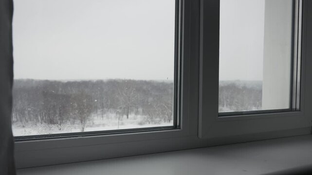 View through white plastic window frame shows snowy park with trees and water drops on glass. Concept of cozy home atmosphere, winter isolation, and cold weather conditions.