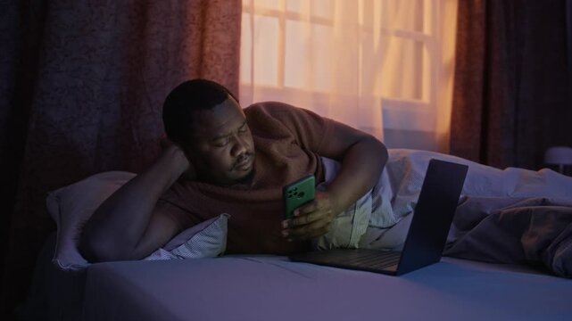 Medium long shot of young African American man lying in bed at night, scrolling on smartphone with laptop open. Video ideal for campaigns on digital addiction, insomnia, remote work lifestyle