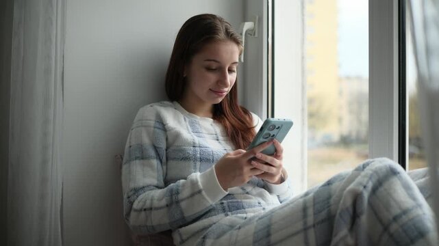 Teenage Girl Sitting On A Windowsill Typing A Message In Social Media On Smartphone
