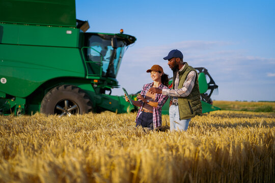 Two farmers using a digital tablet while inspecting a wheat field beside a modern combine harvester during harvest. Diverse agricultural team managing crops with precision farming technology