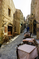 Obraz premium A narrow cobblestone street lined with stone buildings and outdoor cafe seating, with two people walking in the distance.