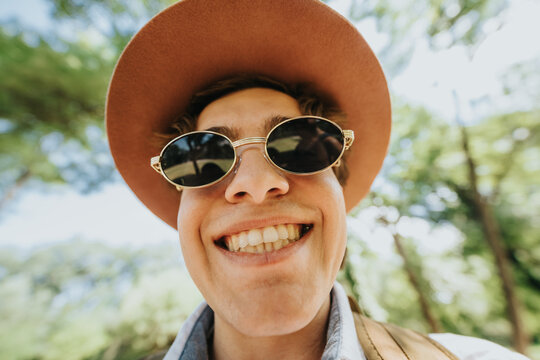 Young man wearing round sunglasses and a wide-brim hat smiles directly at the camera. Close-up outdoor portrait captures a cheerful moment in a sunlit park.