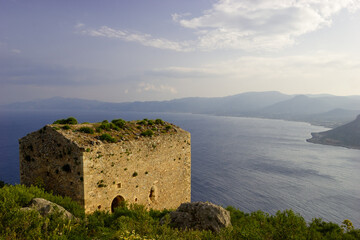 Fototapeta premium A stone tower stands on a grassy hill overlooking the sea and distant mountains under a cloudy sky.