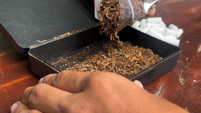 A person pours dried herbs from a clear jar into an open black box on a wooden surface.