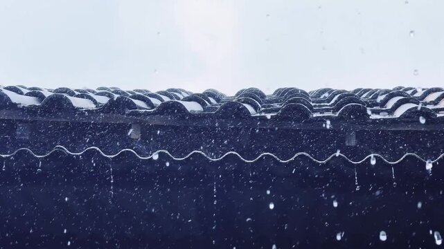 Snow falls gently on a dark roof with rainwater gutter system during stormy weather conditions