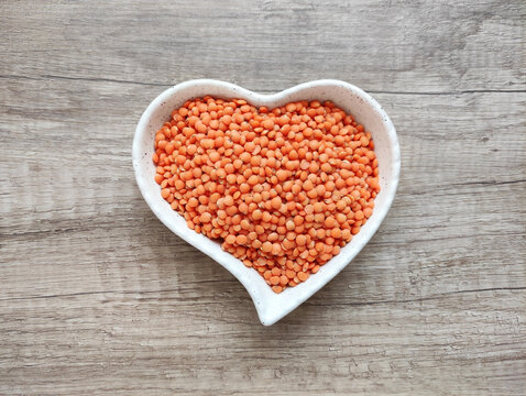 Red lentils in a decorative heart-shaped bowl on a brown wooden background. Healthy plant-based food, nutritious ingredient, top view, soft lighting.