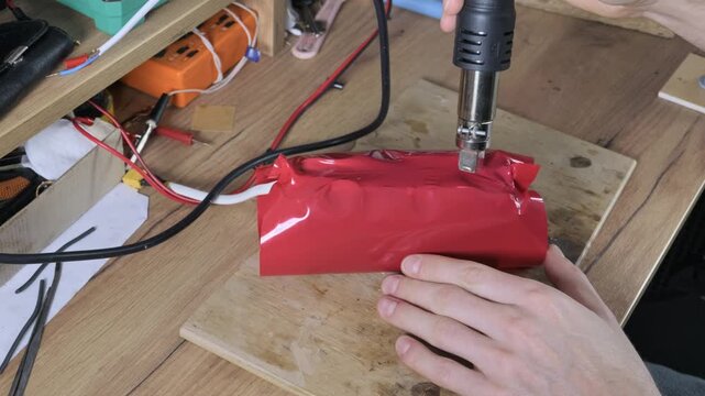 Hands use a heat gun to shrink red PVC tubing around a lithium battery pack on a wooden workbench. This close-up process illustrates precise DIY electronics assembly and insulation.