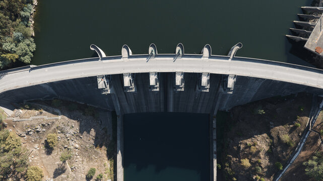 Aerial view of Barragem do Torrao, a concrete dam structure spanning the river with a road on top, surrounded by rocky terrain and dense vegetation in Alpendurada e Matos, Porto, Portugal.