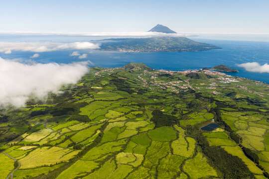 Aerial view of the lush green patchwork fields of the Caldeira region on Faial Island with the volcanic peak of Pico Island rising above the clouds, Horta, Azores, Portugal.