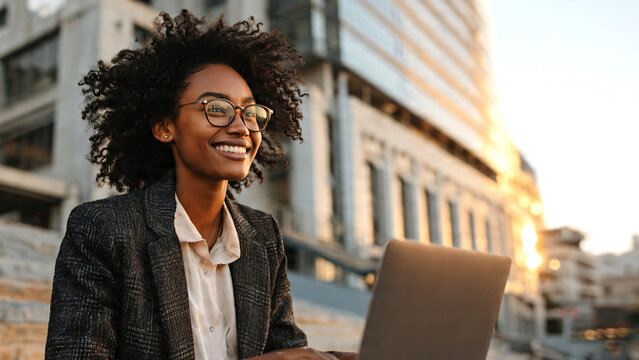 Happy Black businesswoman smiling while working remotely in a modern urban city environment, typing on a laptop computer and creating copywriting content for email marketing campaigns. 