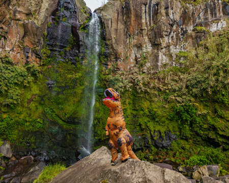 Aerial view of a person in a T-Rex costume standing on a rock before the Cascata das Lombadas waterfall surrounded by lush green cliffs in Conceicao, Acores, Portugal.