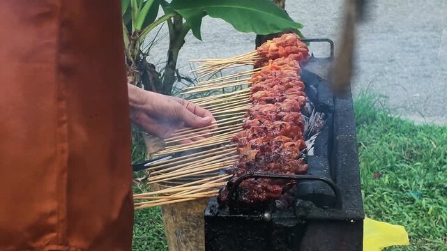 Hands adjust skewers over a small grill. Smoke rises as meat sizzles on bamboo sticks. Grass and leaves frame a rustic outdoor cooking scene. Warm. No animals or specific plants are identifiable