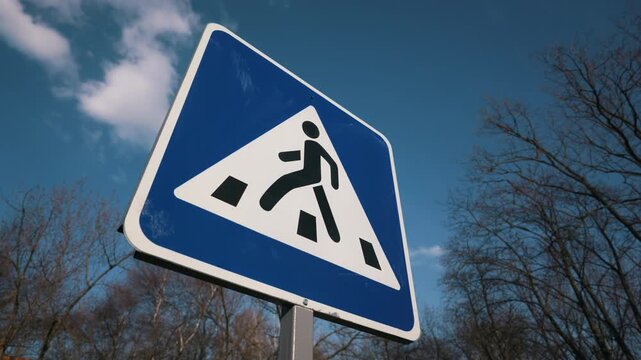 A bright blue pedestrian crossing sign stands prominently against a clear blue sky with light clouds. The low angle perspective emphasizes the traffic symbol among bare winter trees.