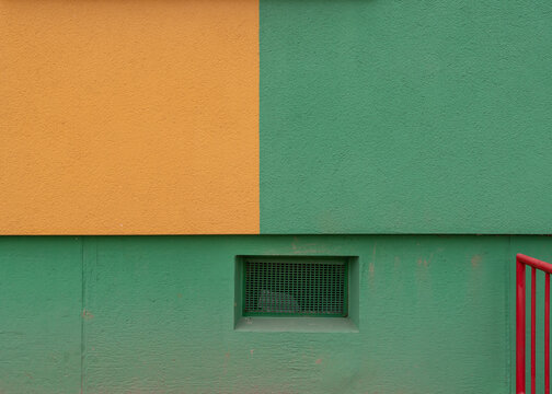 Abstract view of a building facade showing a strict geometric intersection of painted orange and green stucco walls above a green foundation with a metal vent.