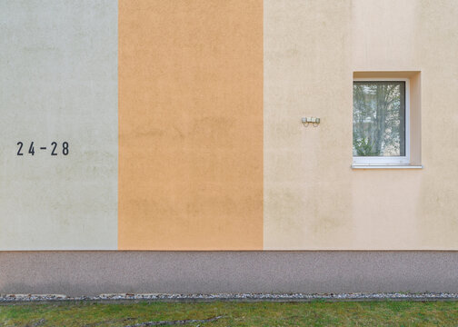 Clean geometric abstract view of a residential building facade showcasing distinct color block sections in sage green, orange, and beige with a single square window.