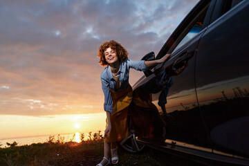Fototapeta premium Smiling woman with curly hair standing beside a car at sunset. Casual denim shirt and dress convey outdoor lifestyle, warmth, energy, and everyday freedom. Natural light creates a bright, candid,