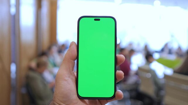 Hand holds phone with green screen. Blurred people sit in background. Ideal for app demos or UI mockups. Clear focus on device for editing. Suitable for business, tech, or presentation projects