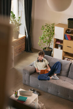 High angle view of teen boy reading book on sofa in modern home. Concept of self education, reading habit, cozy learning, calm lifestyle and green interior.