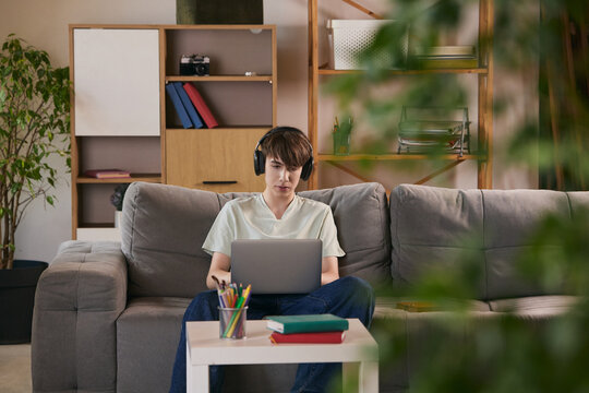 Teen boy studying with laptop on sofa in cozy home learning space. Concept of online education, home study, remote learning, digital skills and modern lifestyle.