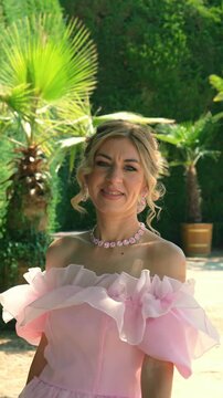 Close-up portrait of a happy Caucasian woman with blonde updo hairstyle, wearing a pink evening dress and matching necklace, smiling directly at the camera in a sunny park.