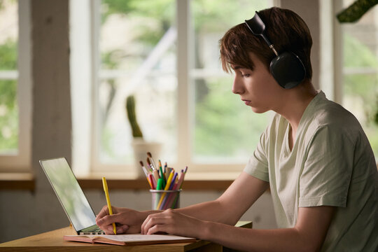 Teen boy taking notes during online study with headphones at desk. Concept of homework routine, remote school, exam preparation, focused learning and productivity.