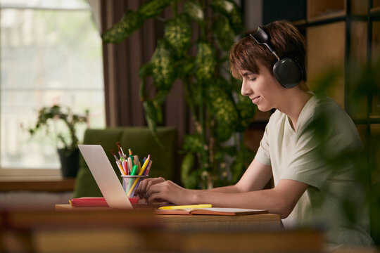 Teen boy studying on laptop with headphones in cozy home workspace. Concept of online learning, home education, remote school, digital skills and productivity.