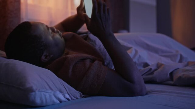 Medium long shot of African American man lying in bed at night, reaching for smartphone and scrolling through screen. Suitable for digital wellness, insomnia