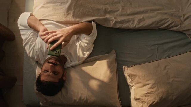 High angle shot of young Indian man waking up in bed, checking smartphone, and turning over to sleep. Perfect for digital detox campaigns or sleep wellness advertisements