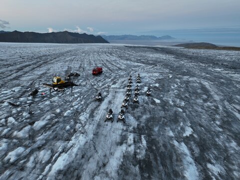 Aerial view of snowmobiles parked on the icy surface of a glacier, with a snowcat vehicle nearby and distant mountains under a twilight sky in Kalfafellsstadur, Hornafjordur, Iceland.
