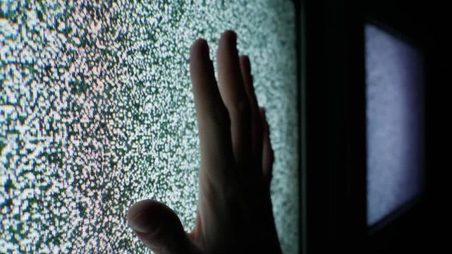 Close-up view of a person hand pressing against a flickering analog television screen displaying white noise static. Concept of nostalgia, digital isolation, and lost media signals.