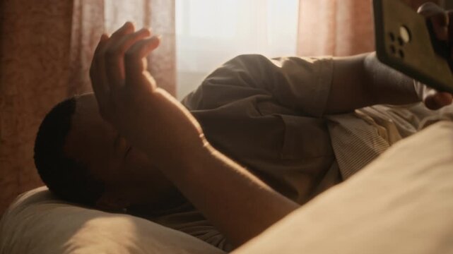 Chest up shot of young African American man waking up in bed, checking smartphone, stretching, and turning over to sleep. Ideal for sleep wellness campaigns, mobile app promotions