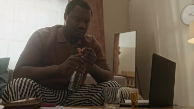 Medium long shot of young Black man pouring water into glass and taking pills from prescription bottle at home. Ideal for pharmaceutical advertisements or mental health awareness