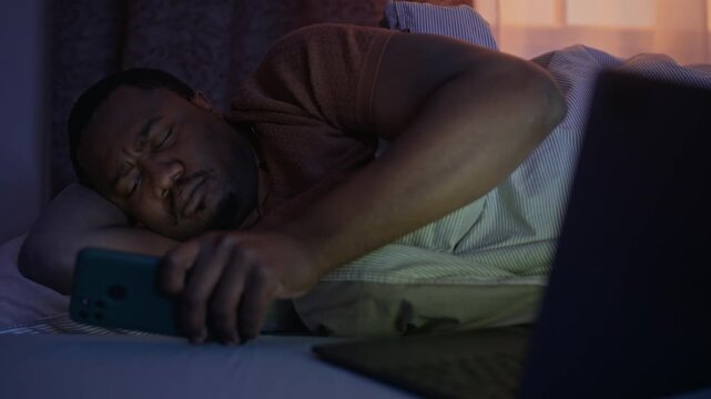 Medium shot of African American young man waking up in bed at night and checking smartphone. Clip is ideal for commercial projects about digital addiction, sleep hygiene, and mobile usage