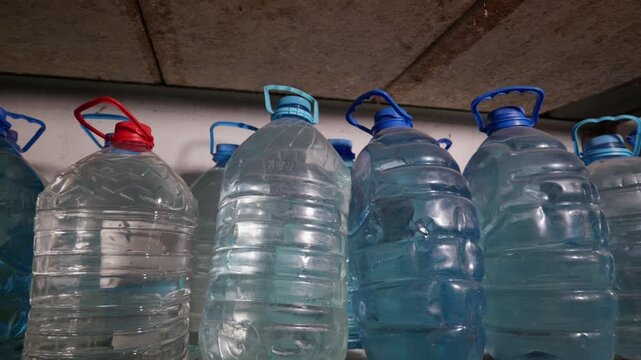 Large plastic bottles filled with fresh drinking water sit securely on a storage shelf. These transparent containers rest in a dimly lit basement to create an emergency supply.