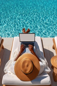 Woman working on a laptop by a sparkling pool while relaxing on a sun lounger with a sun hat